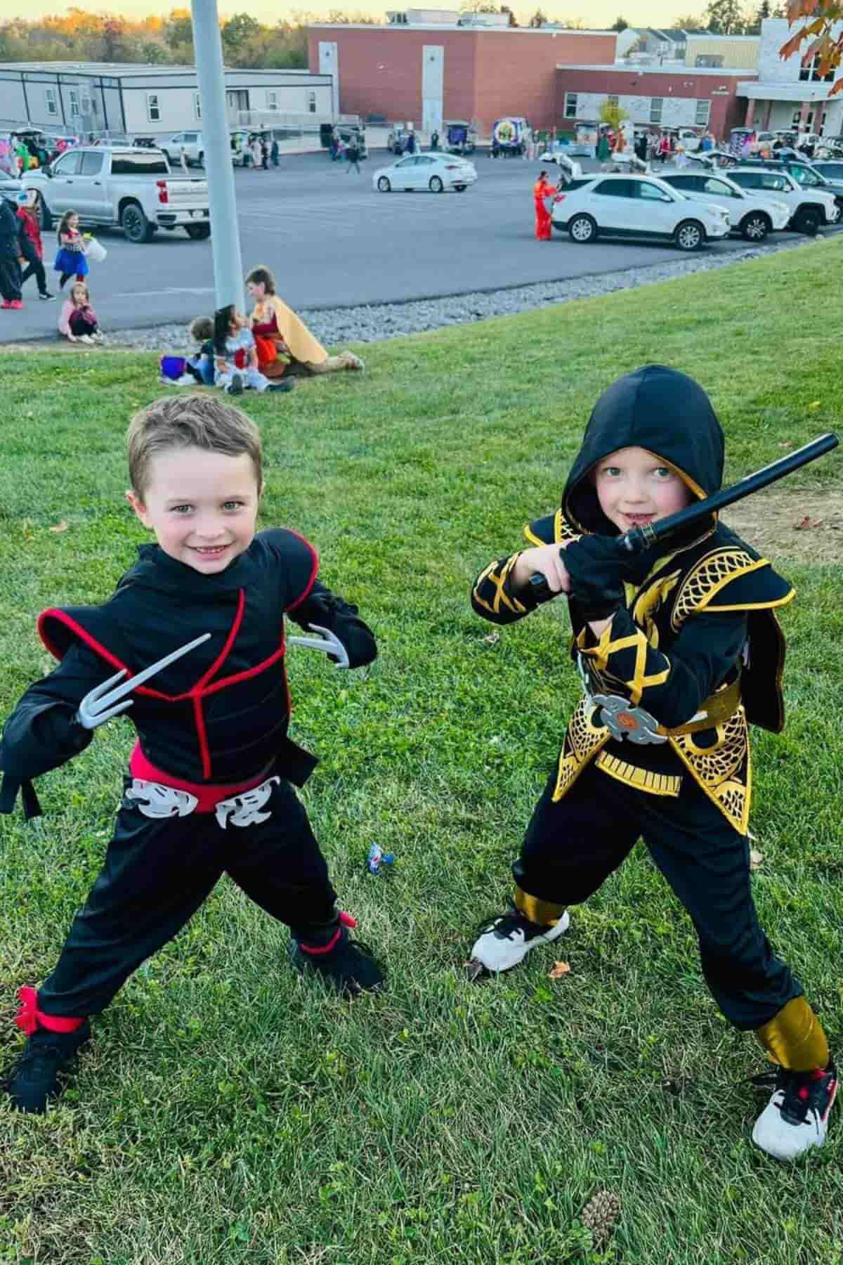 Two children in ninja costumes pose with plastic weapons on a grassy area, with cars and other people in costumes visible in the background.