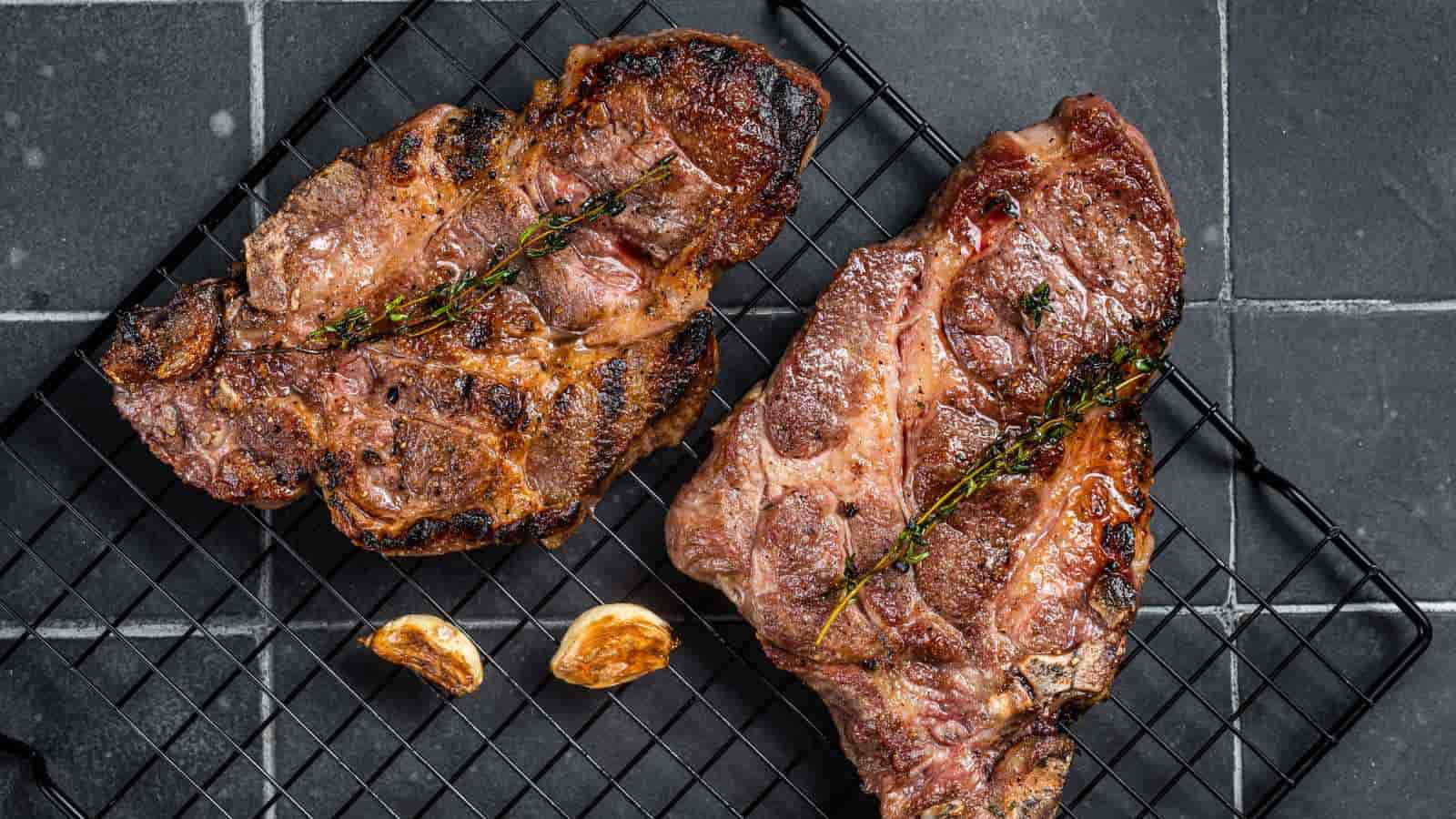 Two cooked steaks garnished with sprigs of herbs rest on a black wire rack with two pieces of roasted garlic, all on a dark tiled surface.
