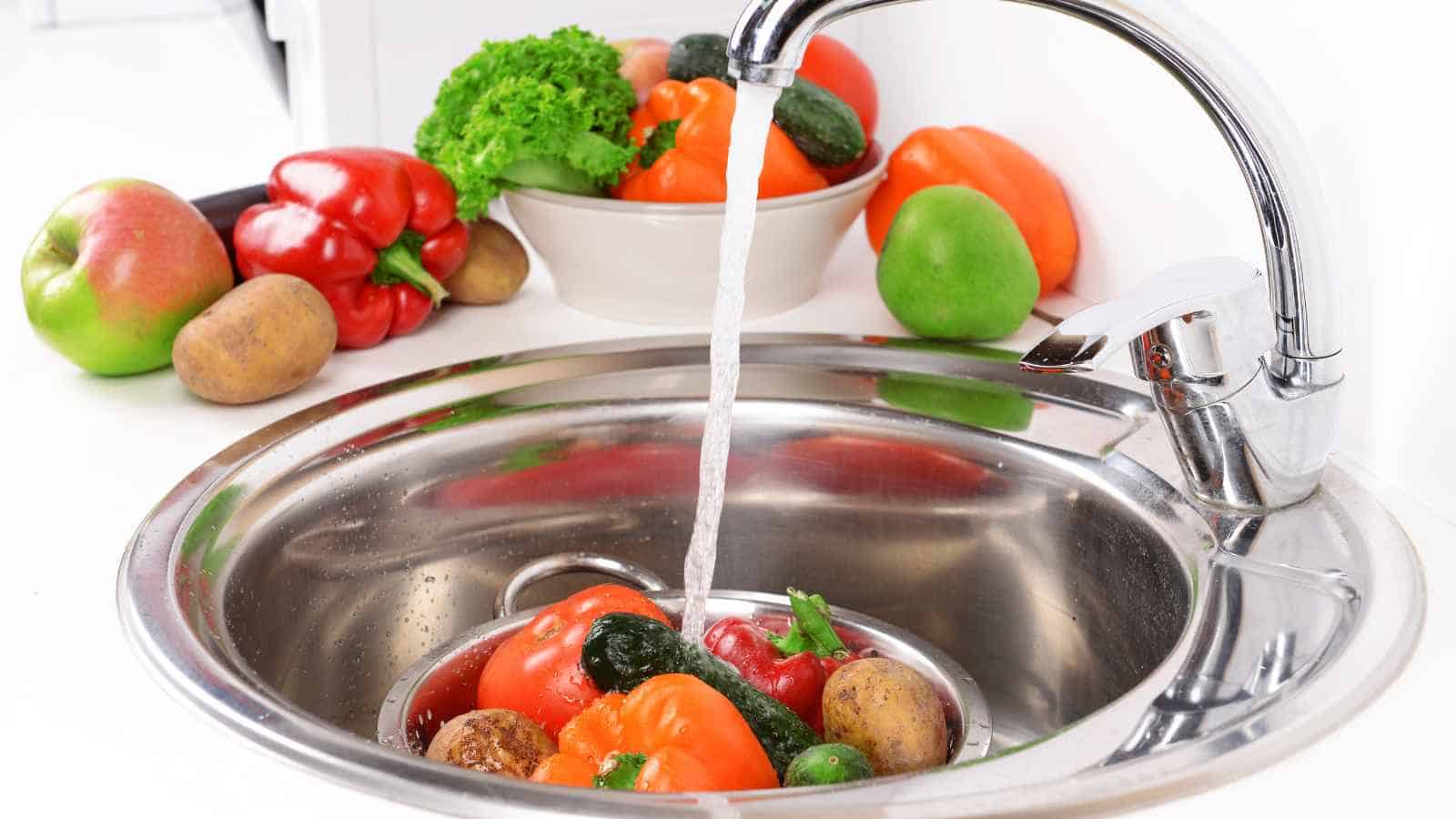 Various fresh vegetables and fruits, including peppers, tomatoes, potatoes, and cucumbers, are being washed under running water in a stainless steel kitchen sink.