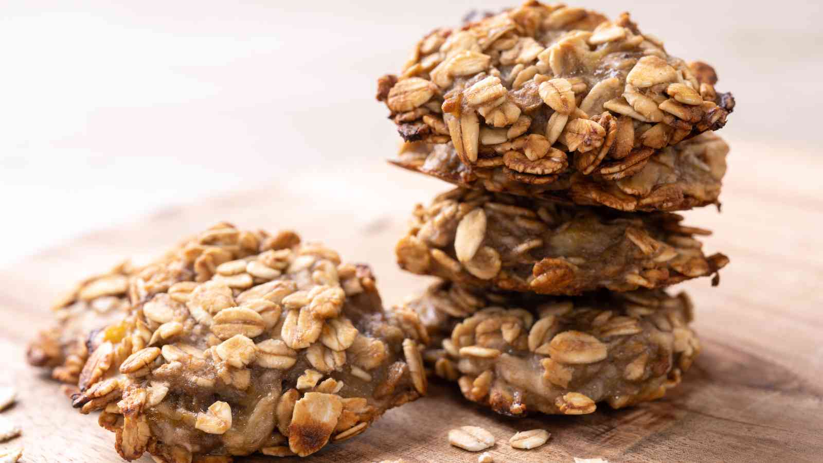 A small stack of oatmeal cookies sits on a wooden surface, with loose oats scattered around.