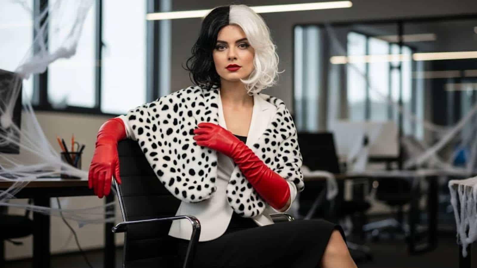 A woman in a black and white wig, spotted shawl, and red gloves sits in an office decorated with fake spider webs—an inspiring idea for women's Halloween costumes for work.