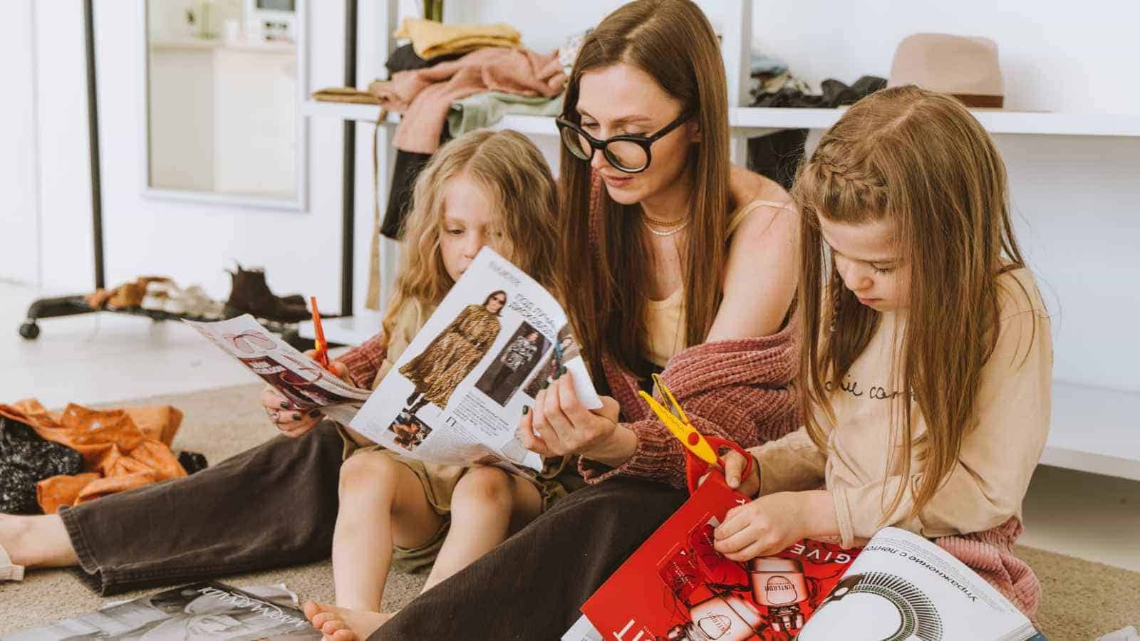 An adult and two children sit on the floor cutting out images from magazines, surrounded by clothing and craft materials.