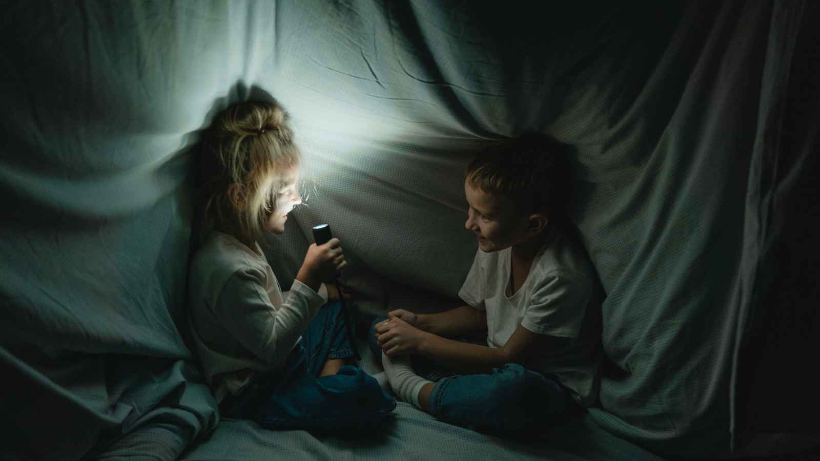 Two children sit inside a blanket fort, one holding a flashlight and speaking, while the other listens and smiles.