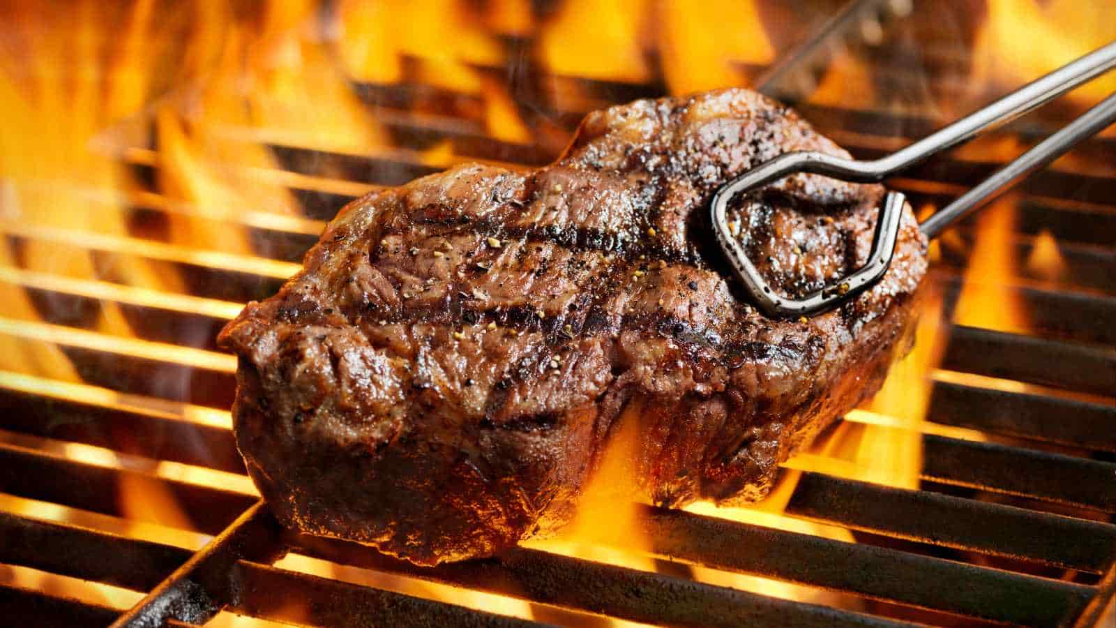 A steak is being grilled over an open flame, with metal tongs holding one end of the meat. Grill marks are visible on the steak.