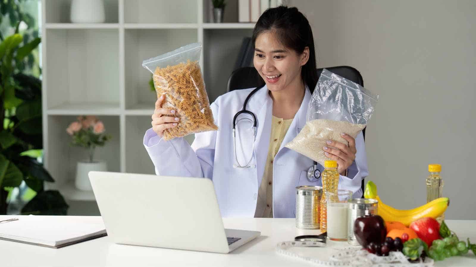 A woman in a lab coat and stethoscope sits at a desk with food items, holding bags of pasta and rice while looking at a laptop.