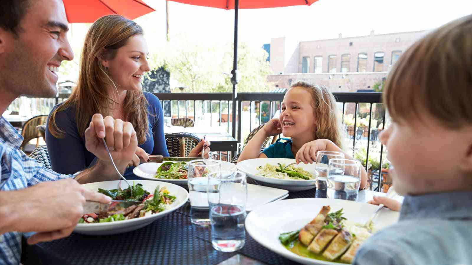 A family of four sits at an outdoor restaurant table, eating salads and smiling, with drinks and plates of food in front of them.