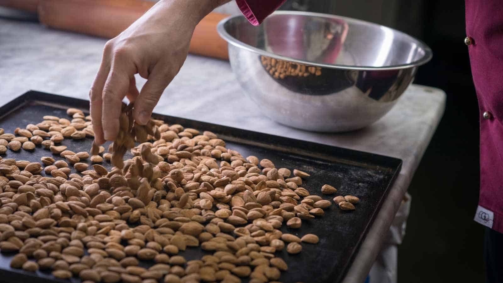 A person’s hand arranging roasted almonds on a baking tray, with a metal mixing bowl in the background on a kitchen counter.