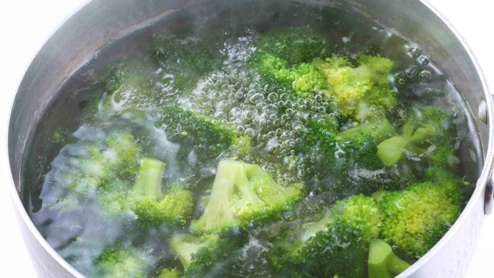Broccoli florets are boiling in a pot of water with visible bubbles and steam rising from the surface.