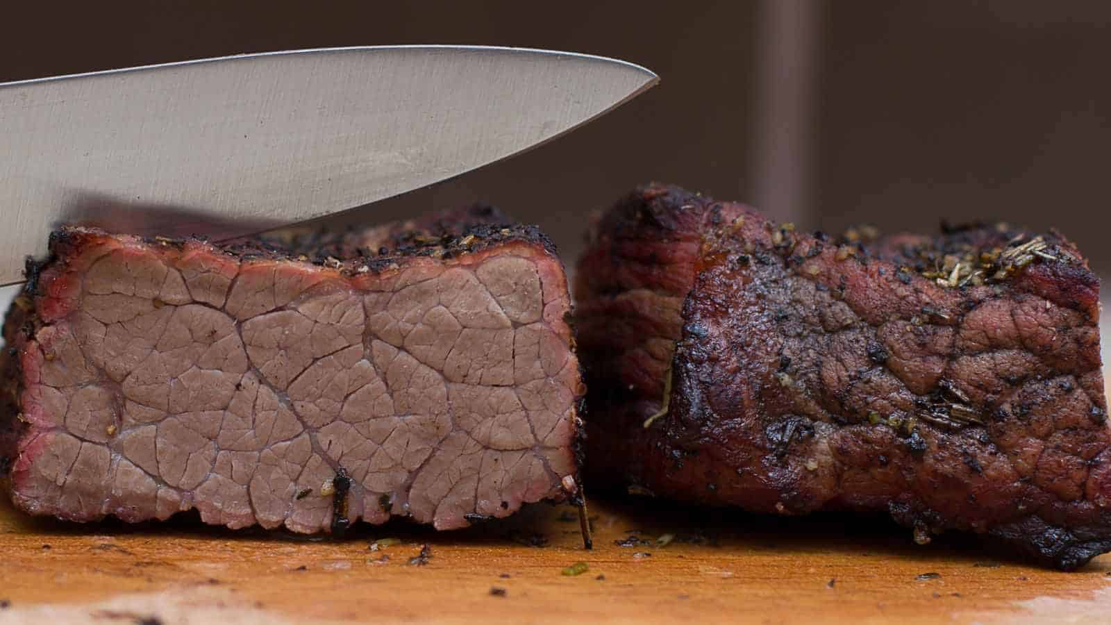 A close-up of a knife slicing through a cooked piece of smoked brisket on a wooden surface.