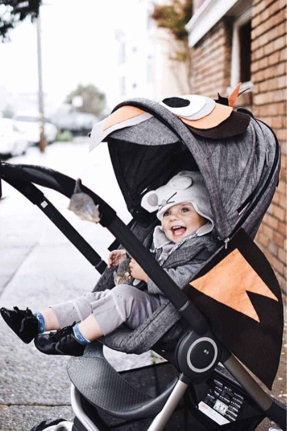 Smiling baby wearing a hooded hat sits in a stroller with a playful owl-themed canopy, outdoors on a city sidewalk by a brick wall.
