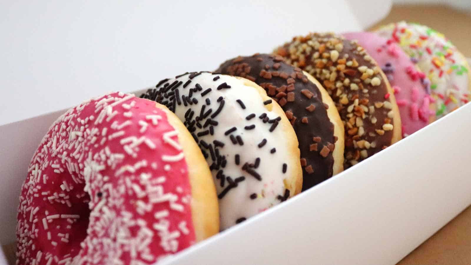 Six assorted donuts with colorful icing and various sprinkles are arranged in a white box.