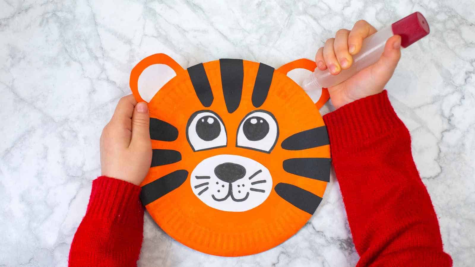 A child in a red sweater holding a paper plate craft shaped and decorated like a tiger's face, placed on a white marble surface.