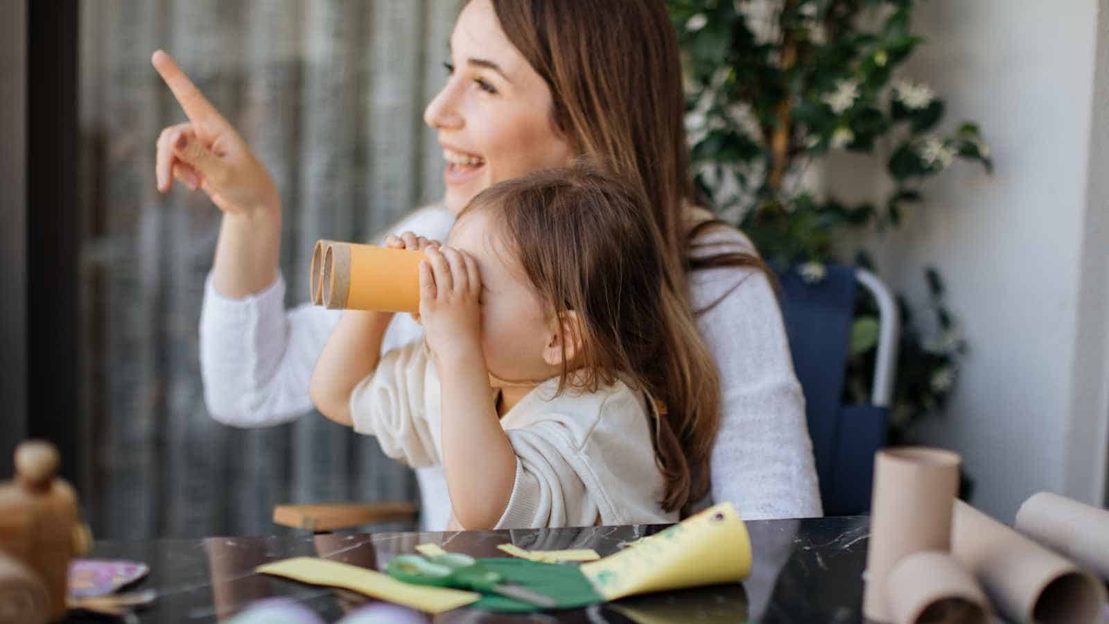 An adult and a child sit at a table with craft supplies; the child looks through a cardboard tube while the adult points into the distance.
