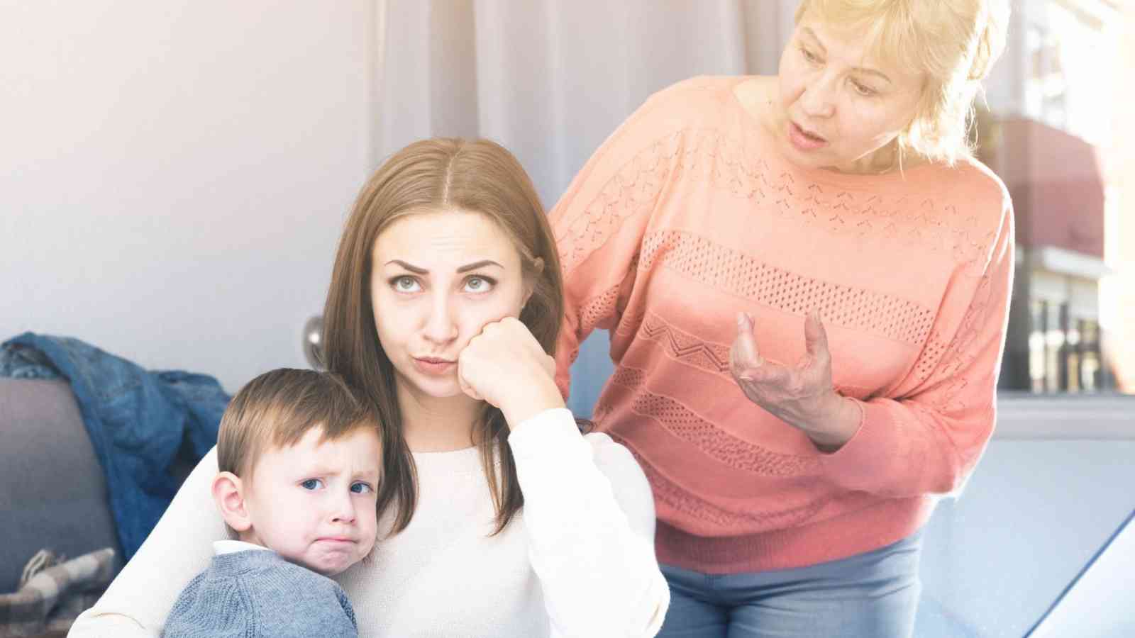 A woman sits holding a sad young boy while an older woman stands behind them, gesturing and appearing to speak to the seated woman.