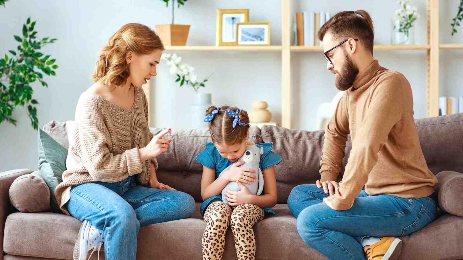 Two adults sit on a couch with a young girl holding a stuffed toy between them; the adults appear to be talking to her, possibly giving advice or scolding.