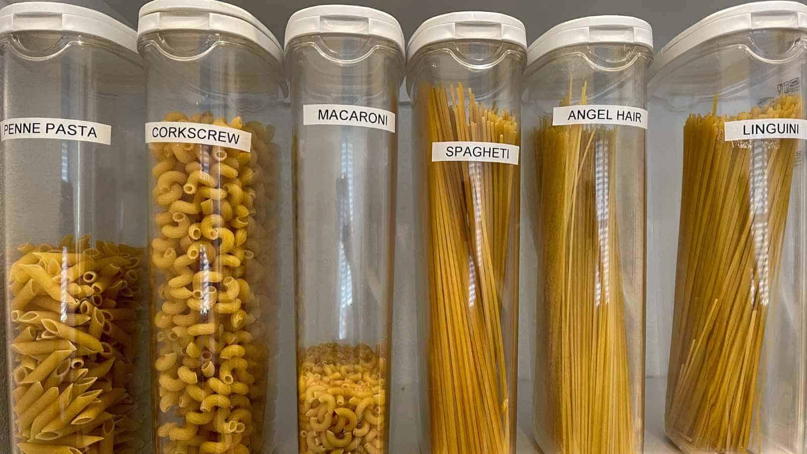 Six clear containers filled with different types of dry pasta: penne, corkscrew, macaroni, spaghetti, angel hair, and linguini, each labeled with its name.