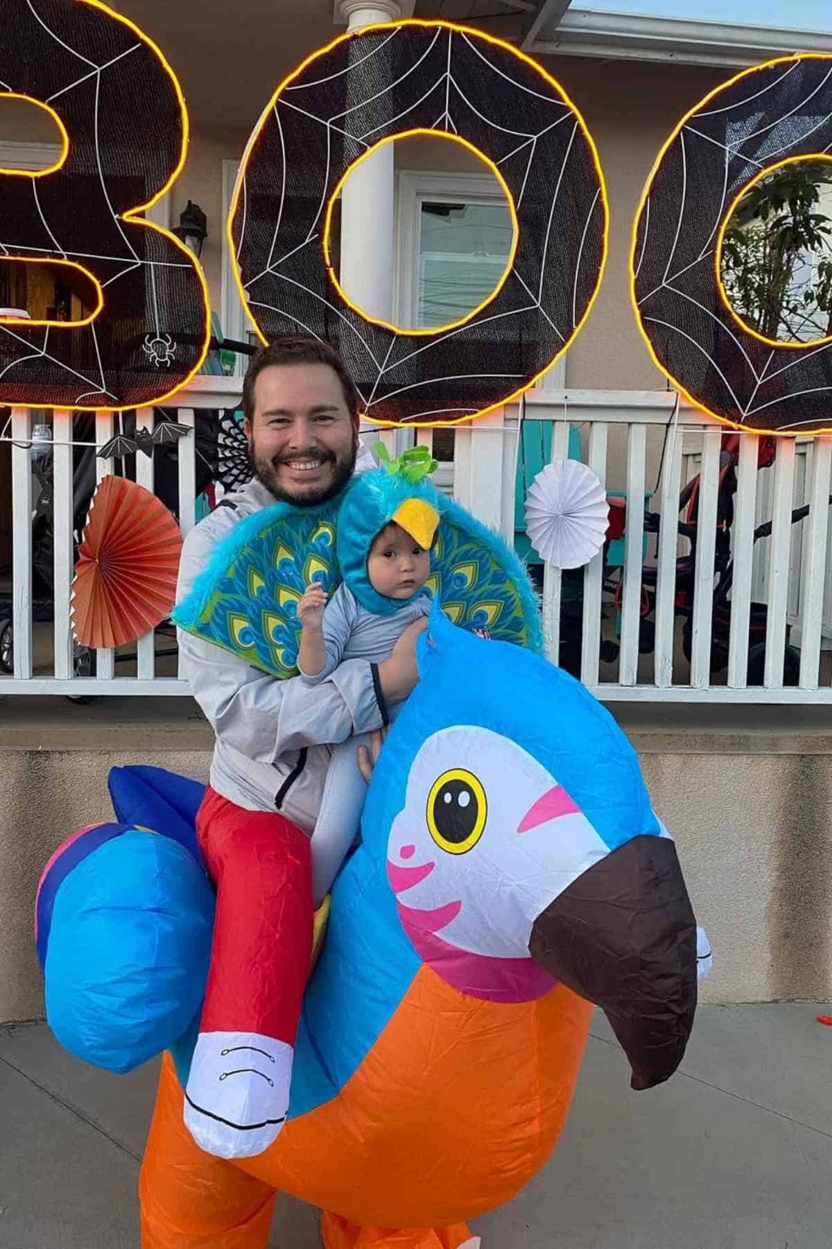 An adult and a baby in parrot and peacock costumes pose in front of a house decorated with spider webs and "BOO" Halloween decorations.