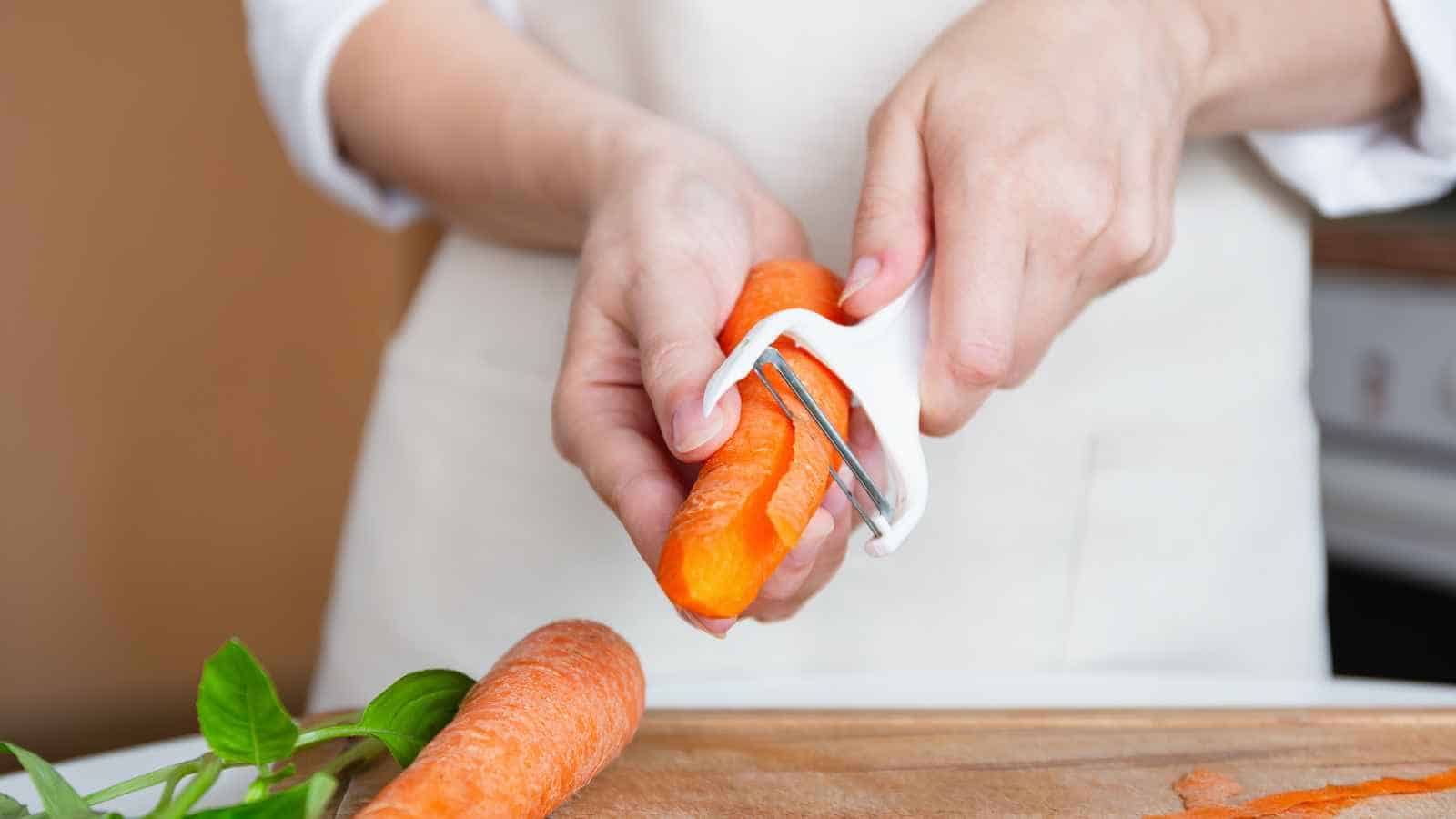 A person uses a white vegetable peeler to peel a carrot over a wooden cutting board.