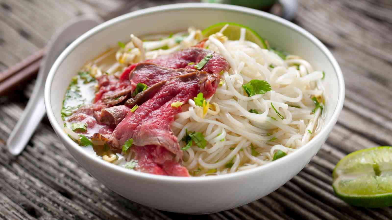 A bowl of pho with rice noodles, thin slices of rare beef, fresh herbs, and lime wedges on a rustic wooden surface.