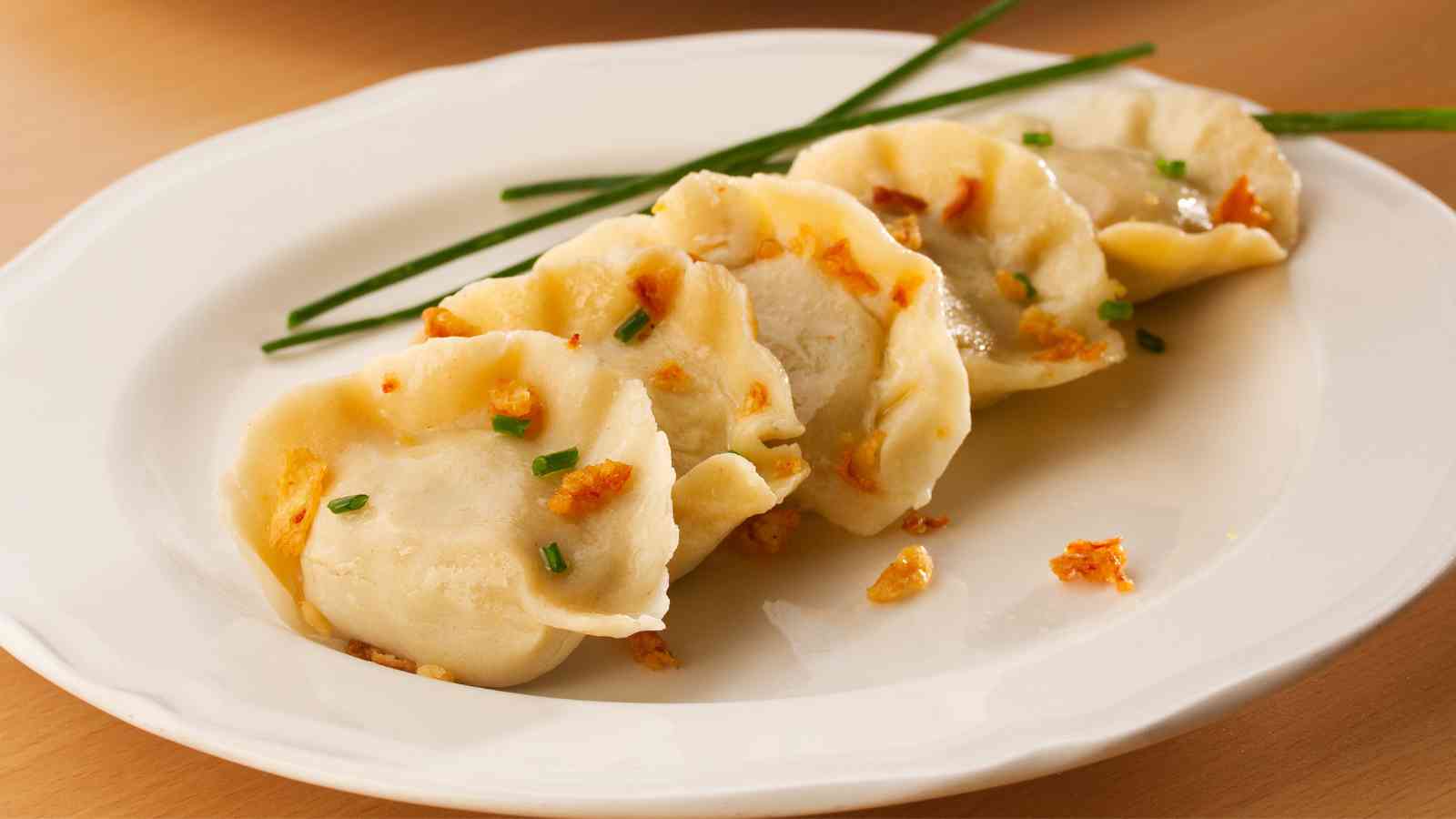 Five pan-fried dumplings garnished with chopped chives and crispy bits are arranged on a white plate, with two whole chives placed diagonally.