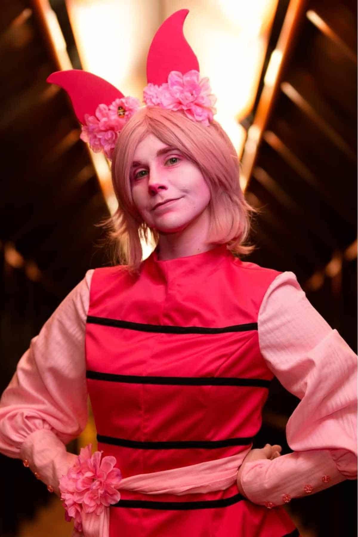 Person wearing a pink costume with flower accents and large pink ear-like headpiece, standing with hands on hips, indoors with warm lighting in the background.