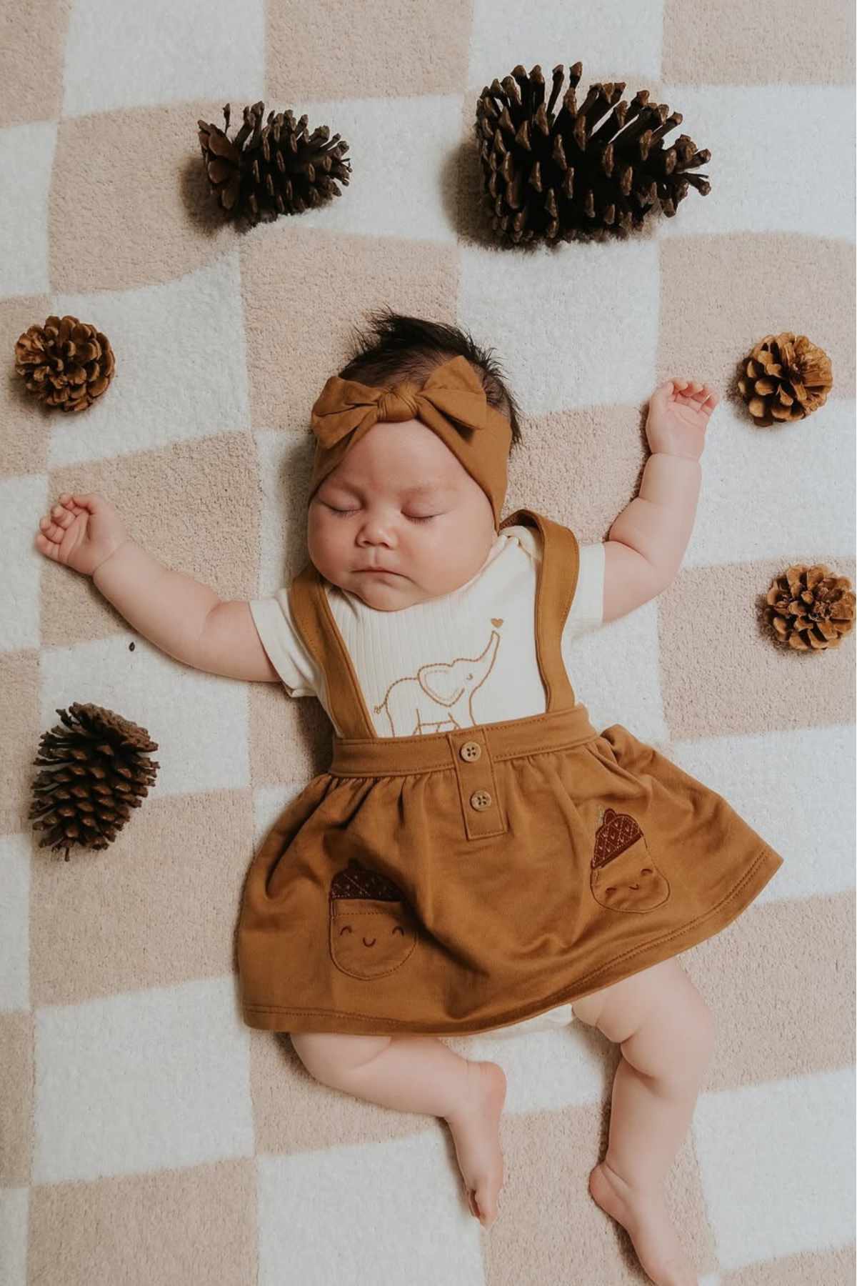 A baby in a brown dress and headband lies on a checkered blanket surrounded by pinecones, arms spread out and eyes closed.
