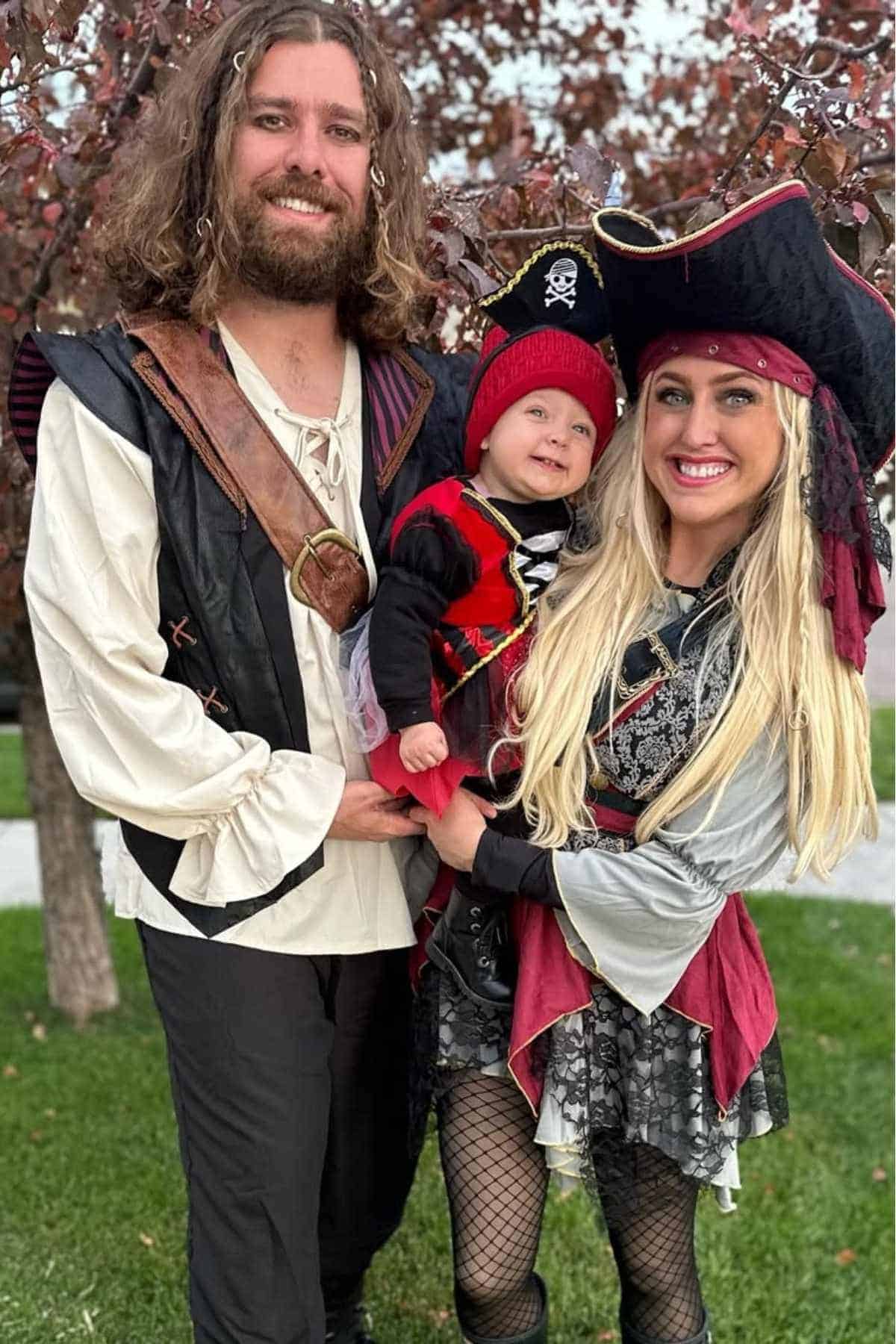 Three people dressed in pirate costumes stand outdoors; two adults smile at the camera while holding a small child between them.