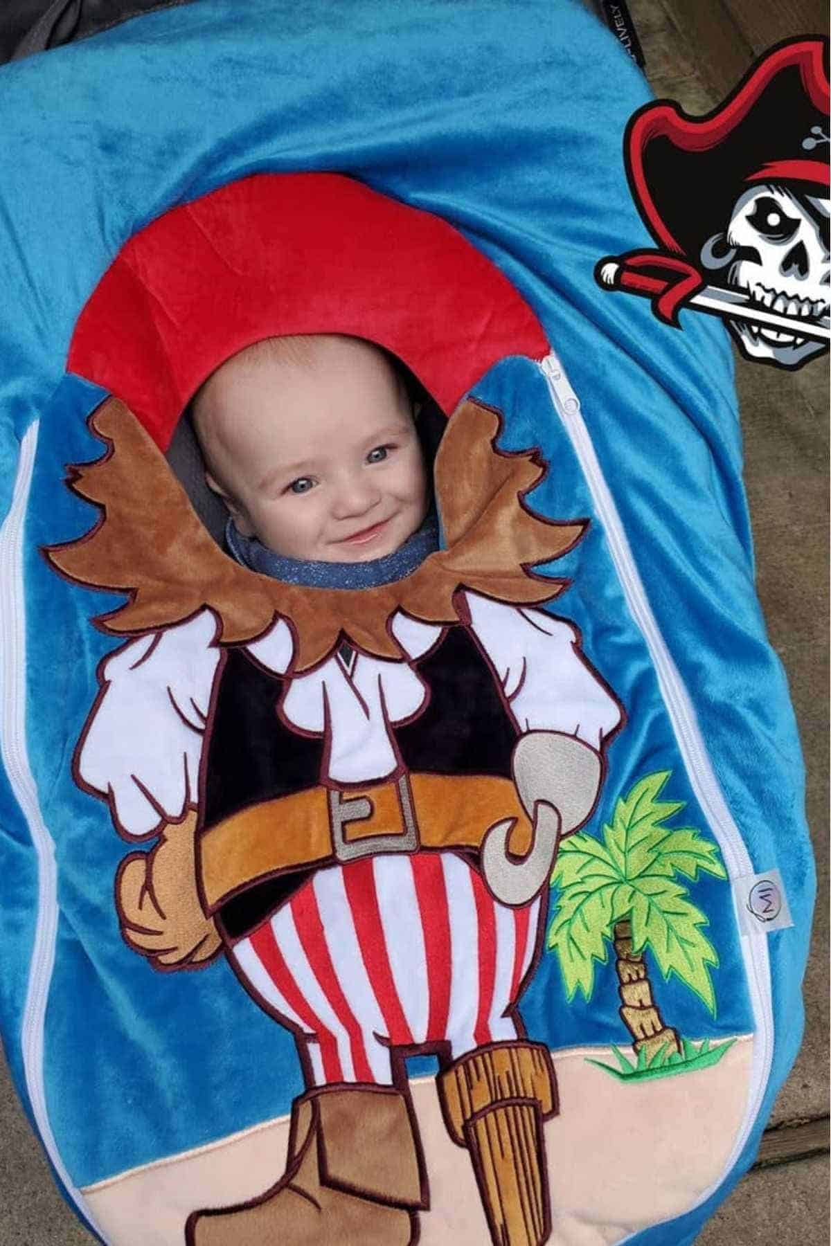 A baby wrapped in a blanket designed to look like a pirate costume, with a pirate hat, striped pants, hook hand, and a palm tree on the beach.