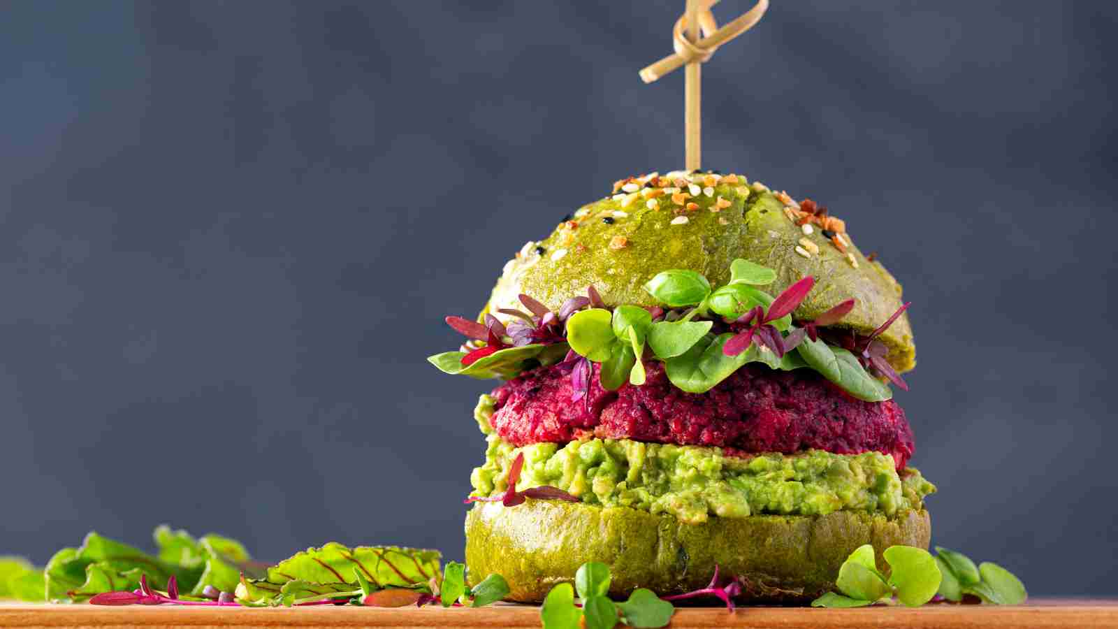 A veggie burger with a green bun, beet patty, guacamole, and mixed greens, garnished with microgreens and held together with a wooden skewer.