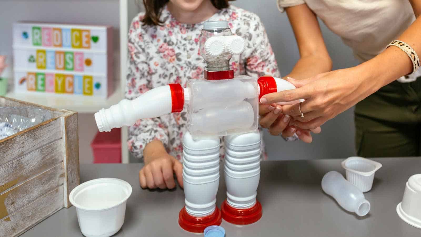 Two people assemble a robot figure using recycled plastic bottles and containers on a table, with a "Reduce Reuse Recycle" sign in the background.
