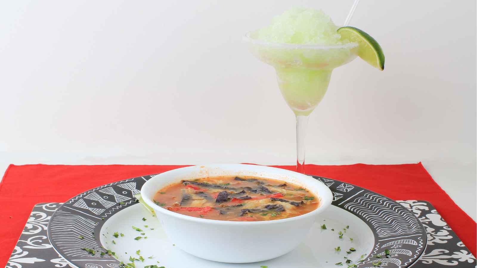 A white bowl of vegetable soup sits on a patterned plate beside a green frozen drink in a margarita glass, garnished with a lime wedge.