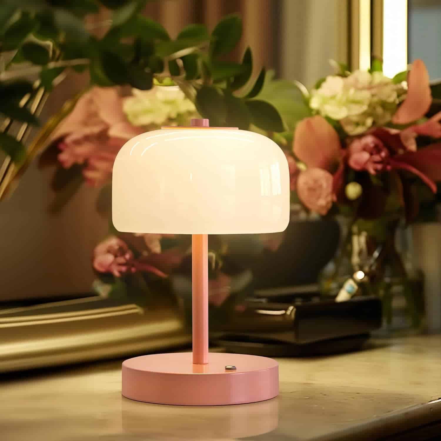 A small pink table lamp with a white dome-shaped shade sits on a marble surface, surrounded by flowers and greenery in the background.