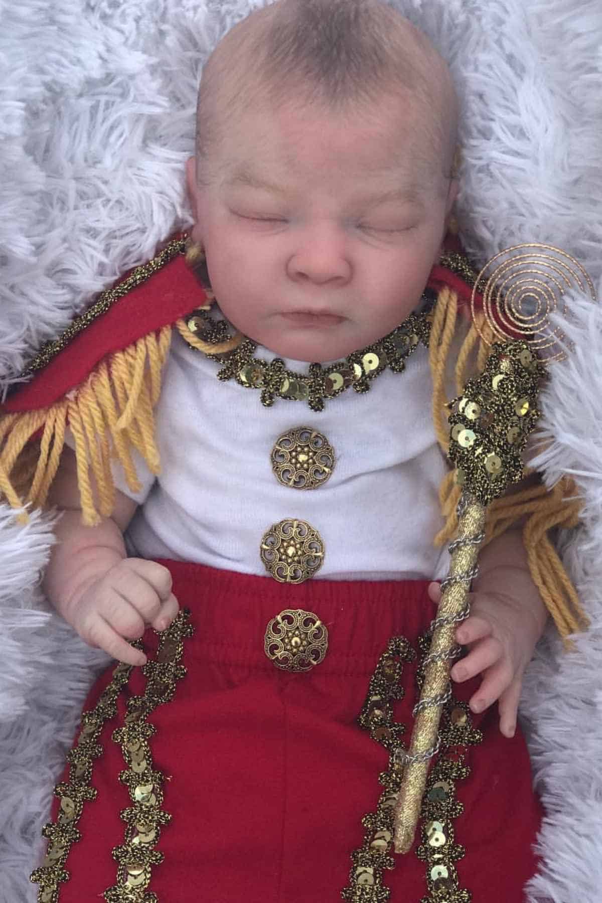 A baby dressed in a decorative red and white costume with gold trim and holding a gold staff, lying on a soft, furry white blanket.