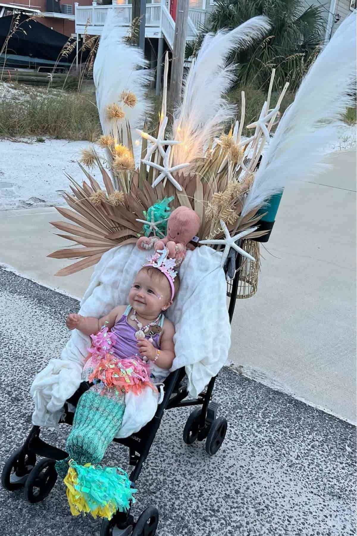 A baby in a stroller are dressed in mermaid costumes with decorative dried plants, starfish, and feathers attached to the stroller.