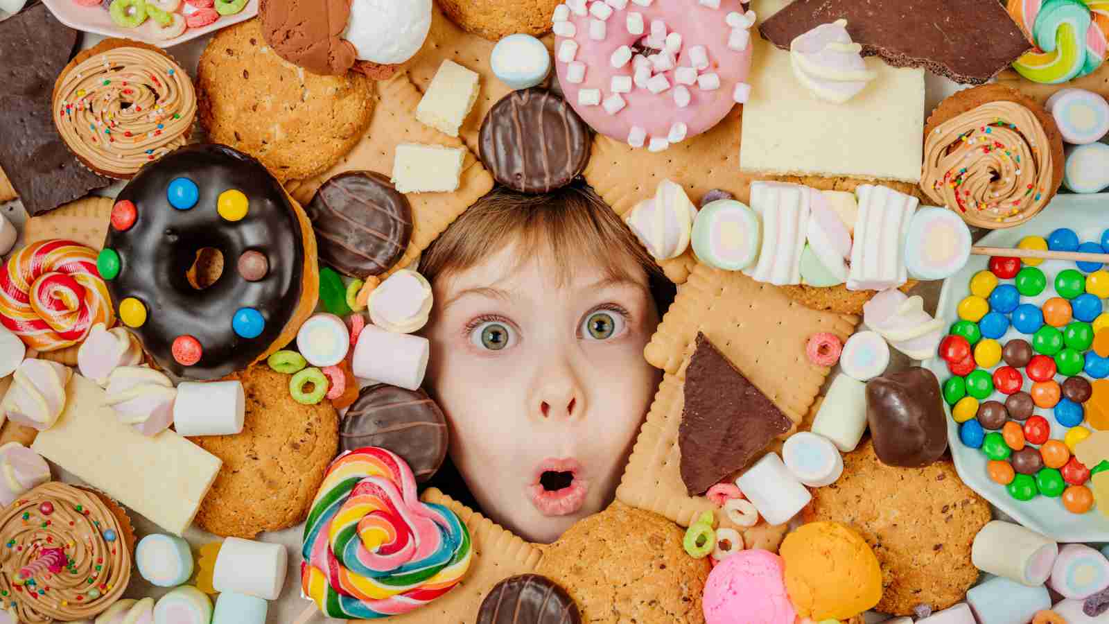 A child’s surprised face peeks through a large assortment of colorful cookies, donuts, candies, and sweets arranged closely together.