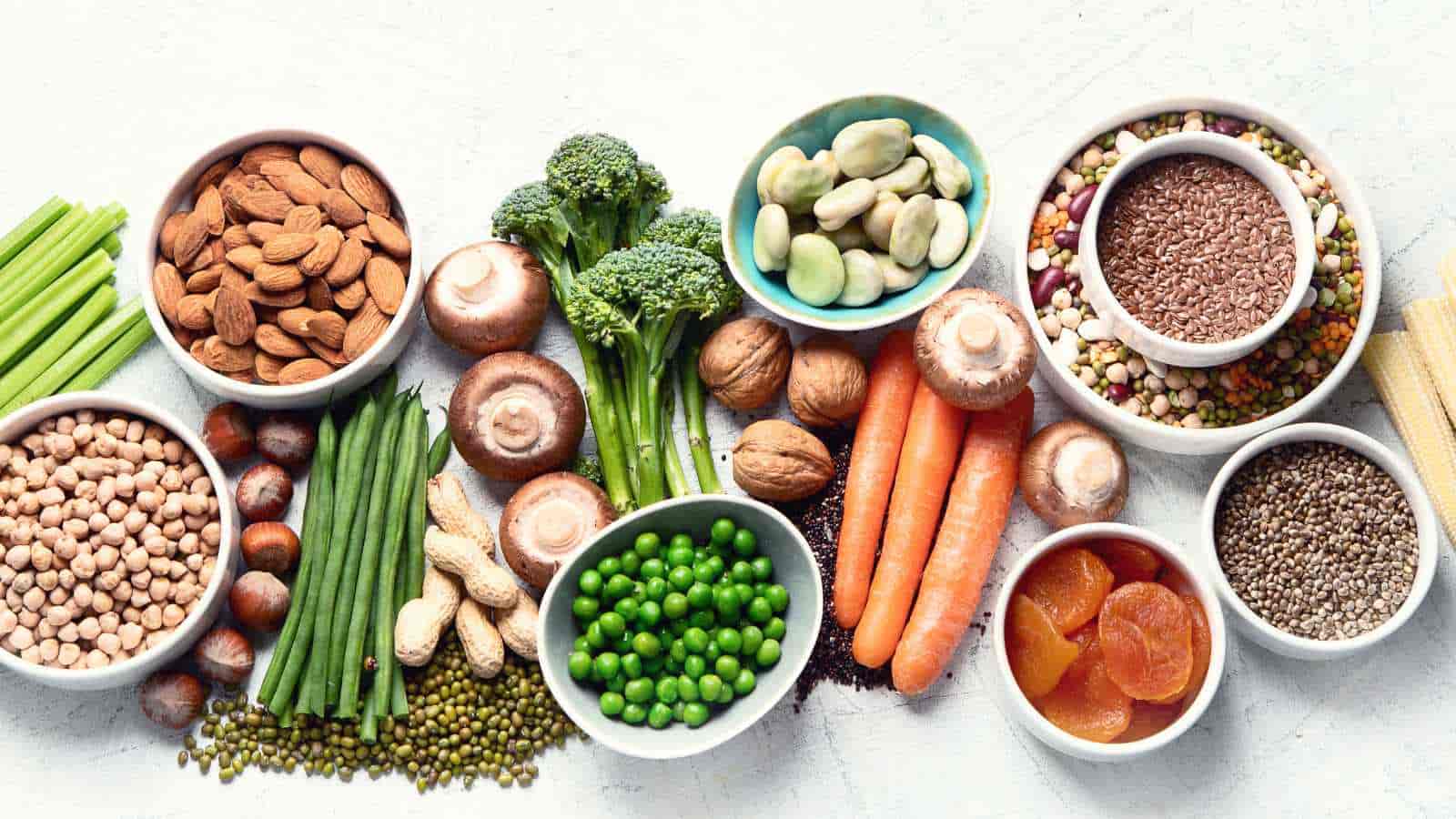 Assorted bowls and piles of vegetables, nuts, seeds, beans, and dried fruit arranged on a white surface.