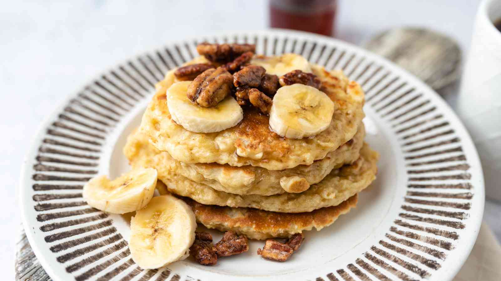 A stack of pancakes topped with banana slices and candied pecans is served on a white plate with a striped rim.