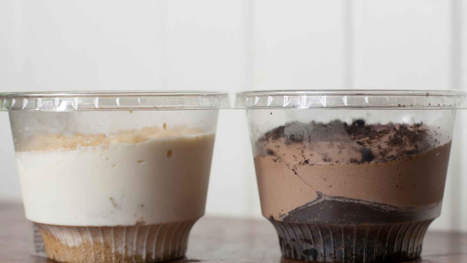 Two clear plastic cups, one filled with a layered vanilla dessert and the other with a layered chocolate dessert, are placed side by side on a wooden surface.