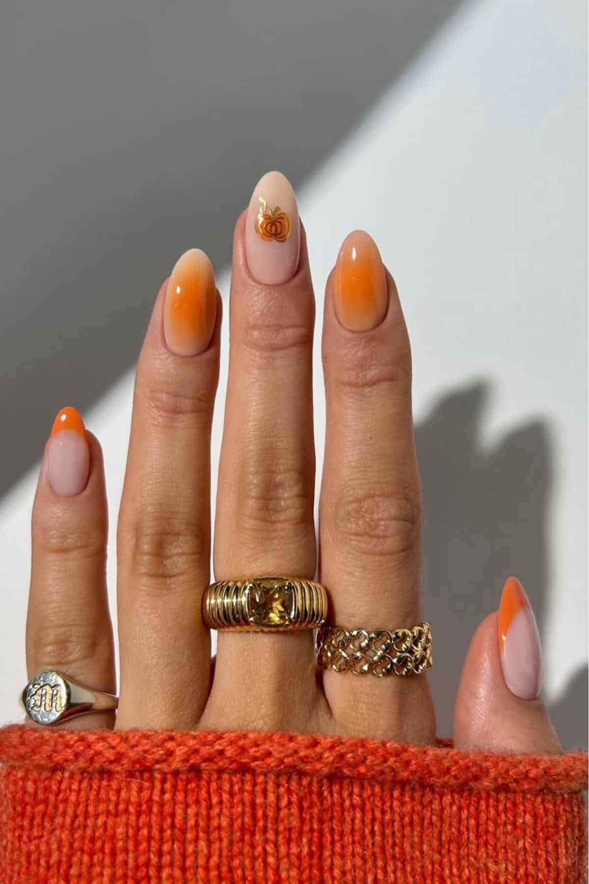 A hand with orange ombre manicured nails, one featuring a small pumpkin design, wears three gold rings and an orange sweater sleeve is partially visible.