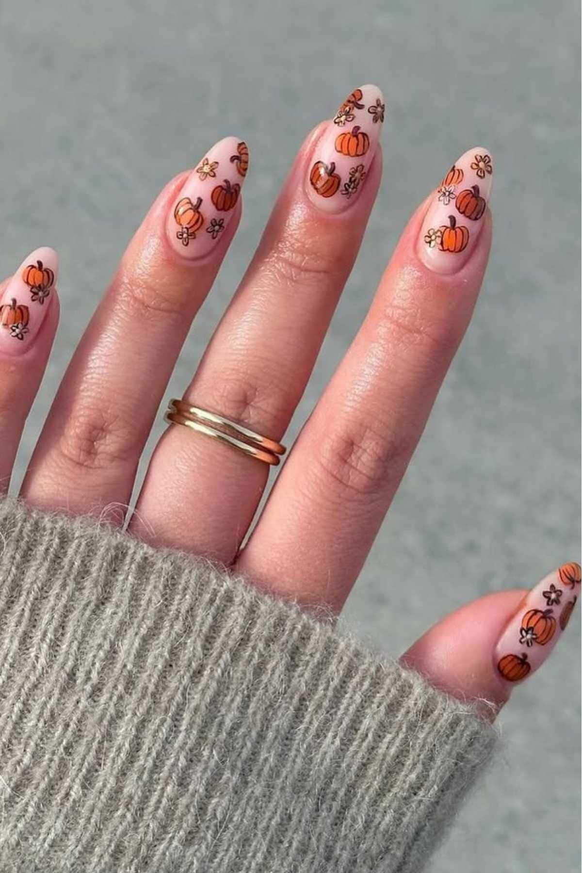 A hand with long almond-shaped nails featuring small orange pumpkin and flower nail art designs, wearing a gold ring and a gray knit sweater.