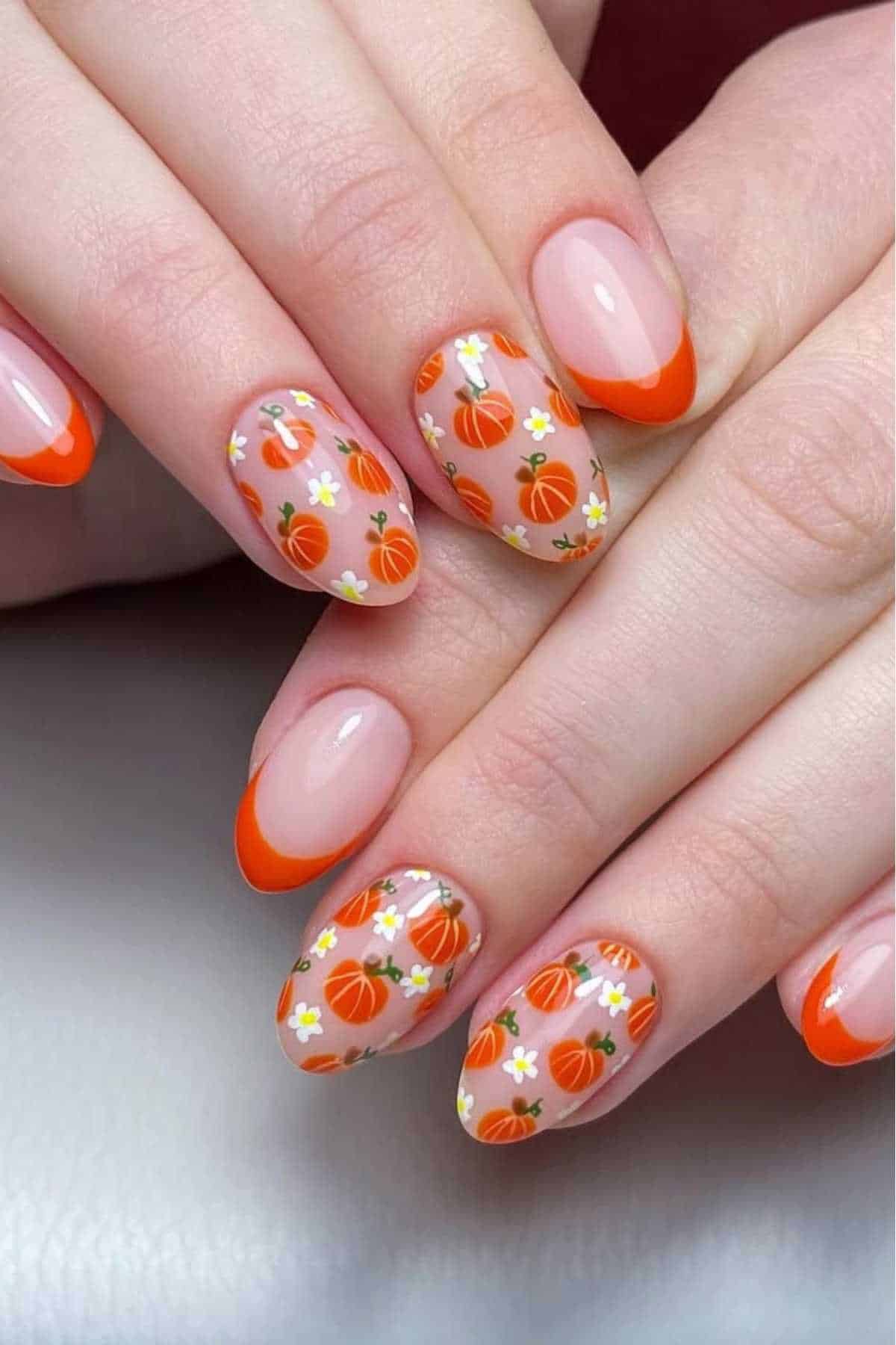Hands with almond-shaped nails featuring orange French tips and nail art with small pumpkins and white flowers on a neutral base.