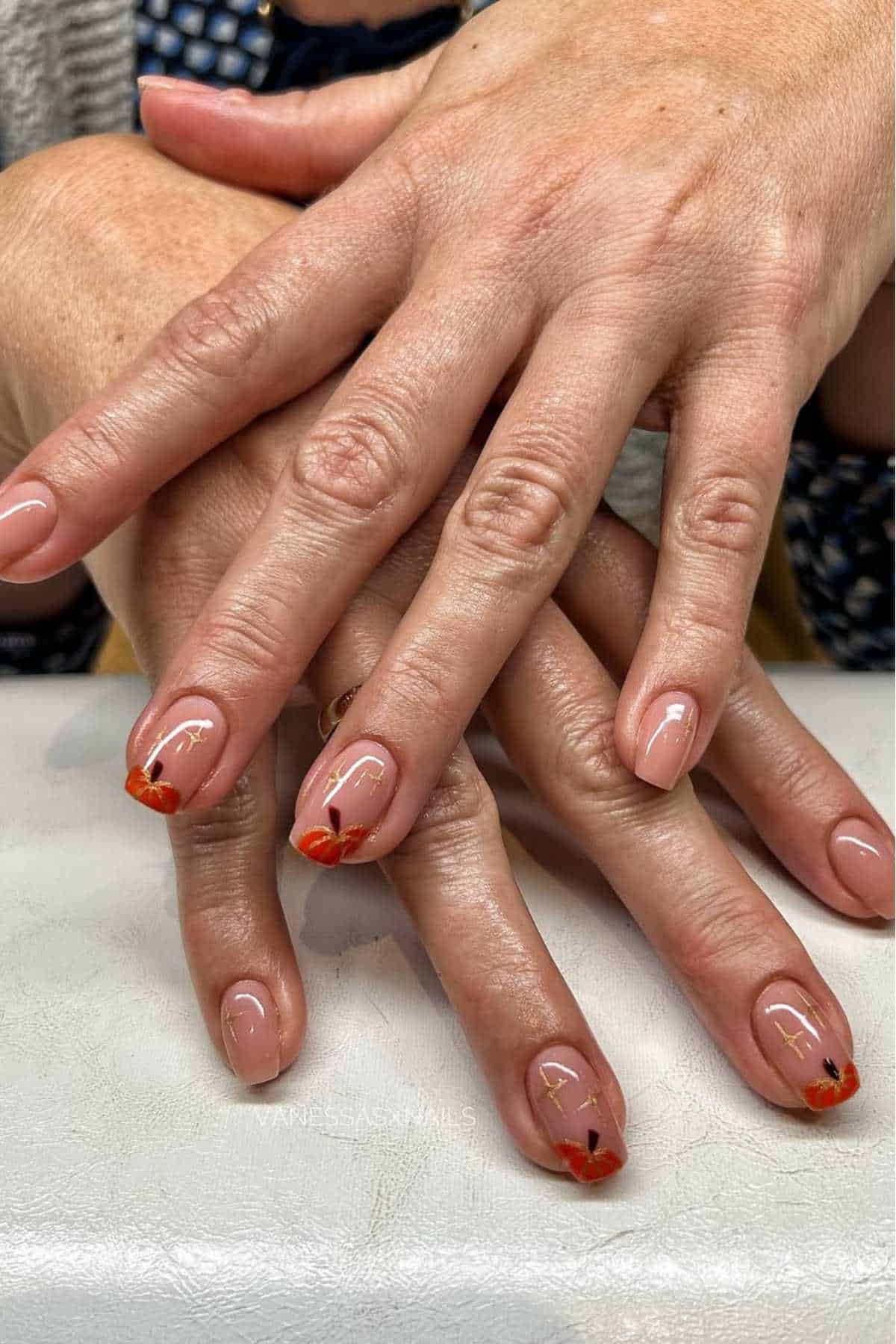 Hands with neatly manicured nails featuring a nude base and small orange and gold abstract designs on the tips, resting on a white surface.
