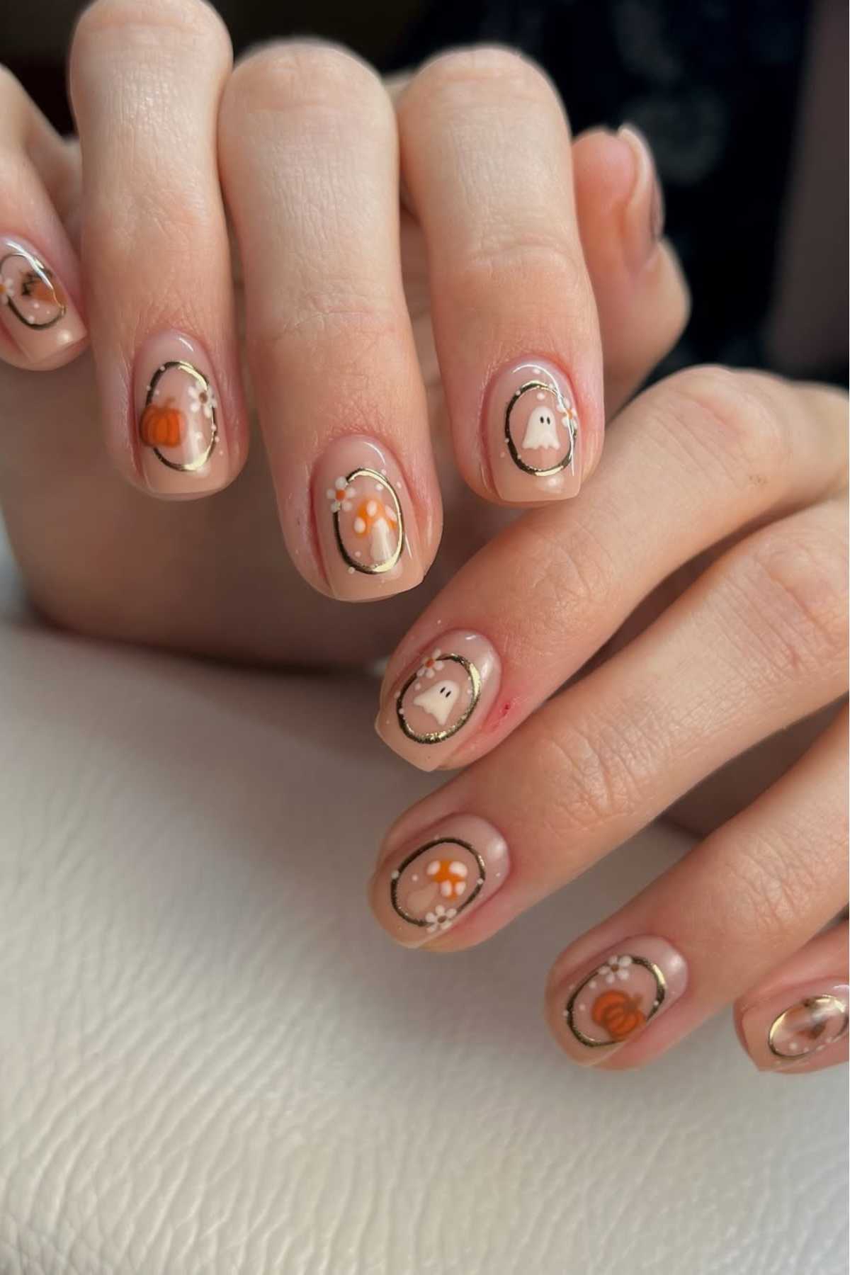 A hand with manicured nails featuring fall-themed nail art, including pumpkins, flowers, and ghost designs on a nude base.