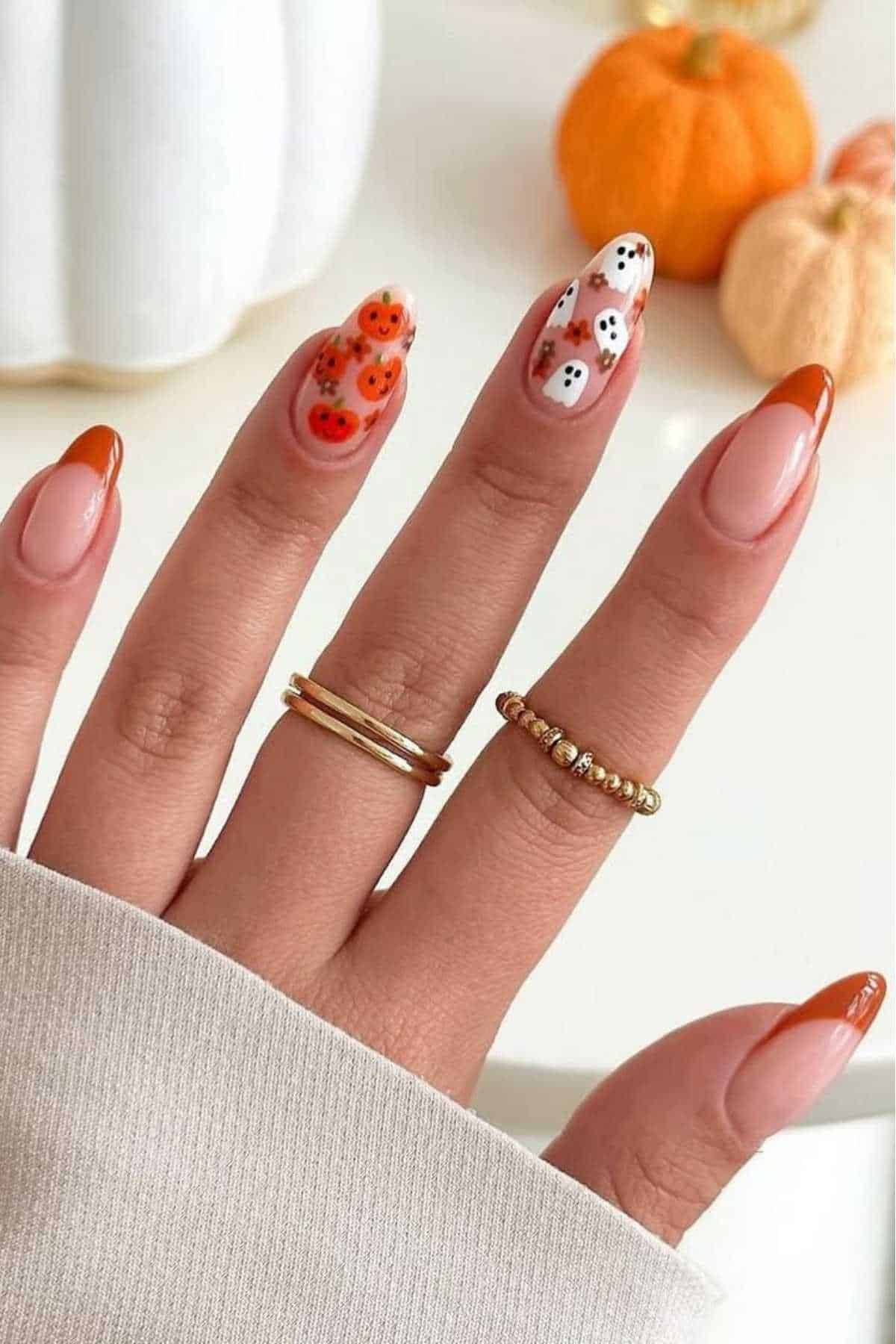 A hand with Halloween-themed nail art, featuring orange French tips, pumpkin designs, and ghost designs, with gold rings and mini pumpkins in the background.
