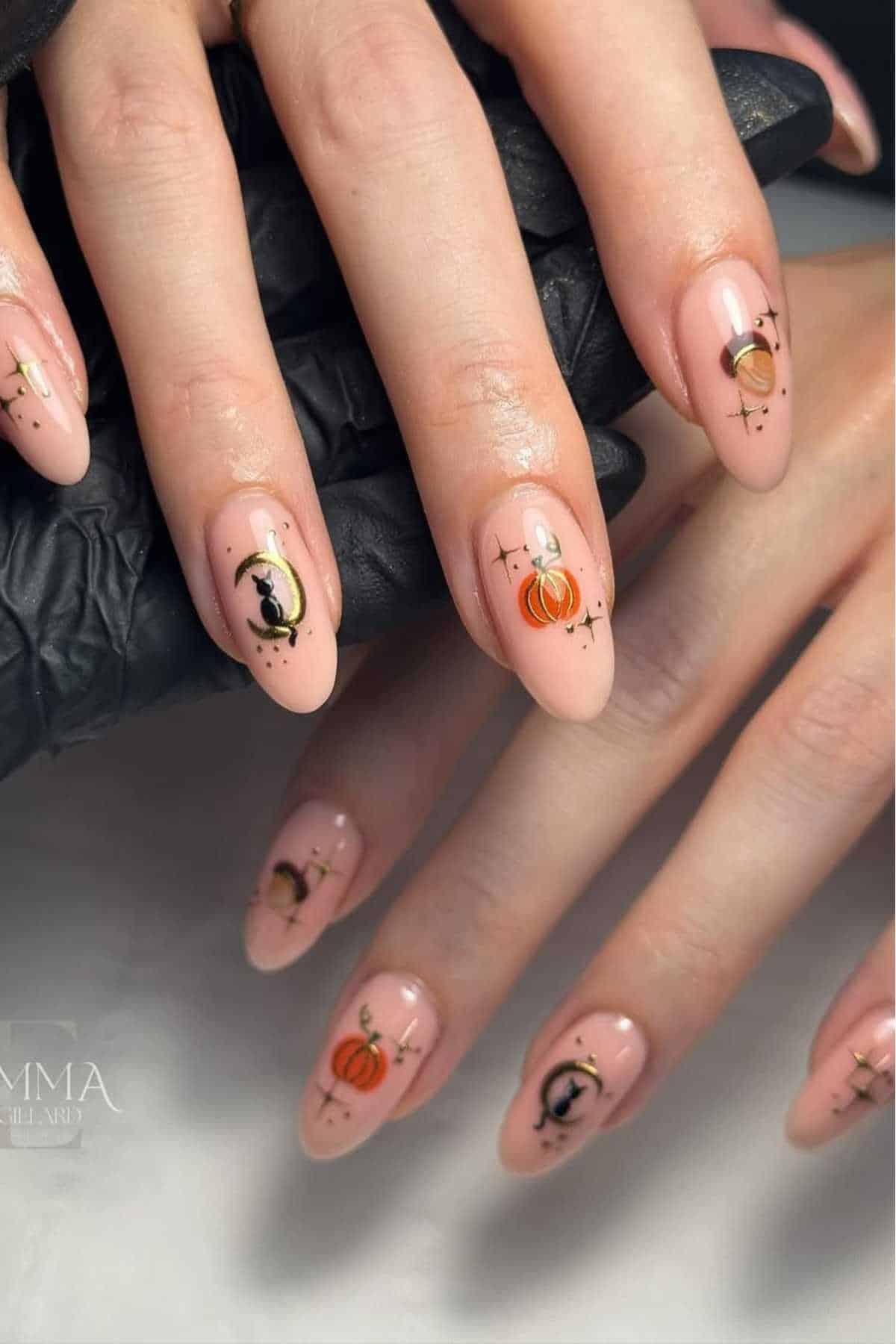 A hand with almond-shaped nude nails featuring Halloween-themed nail art, including pumpkins, black cats, moons, and stars.