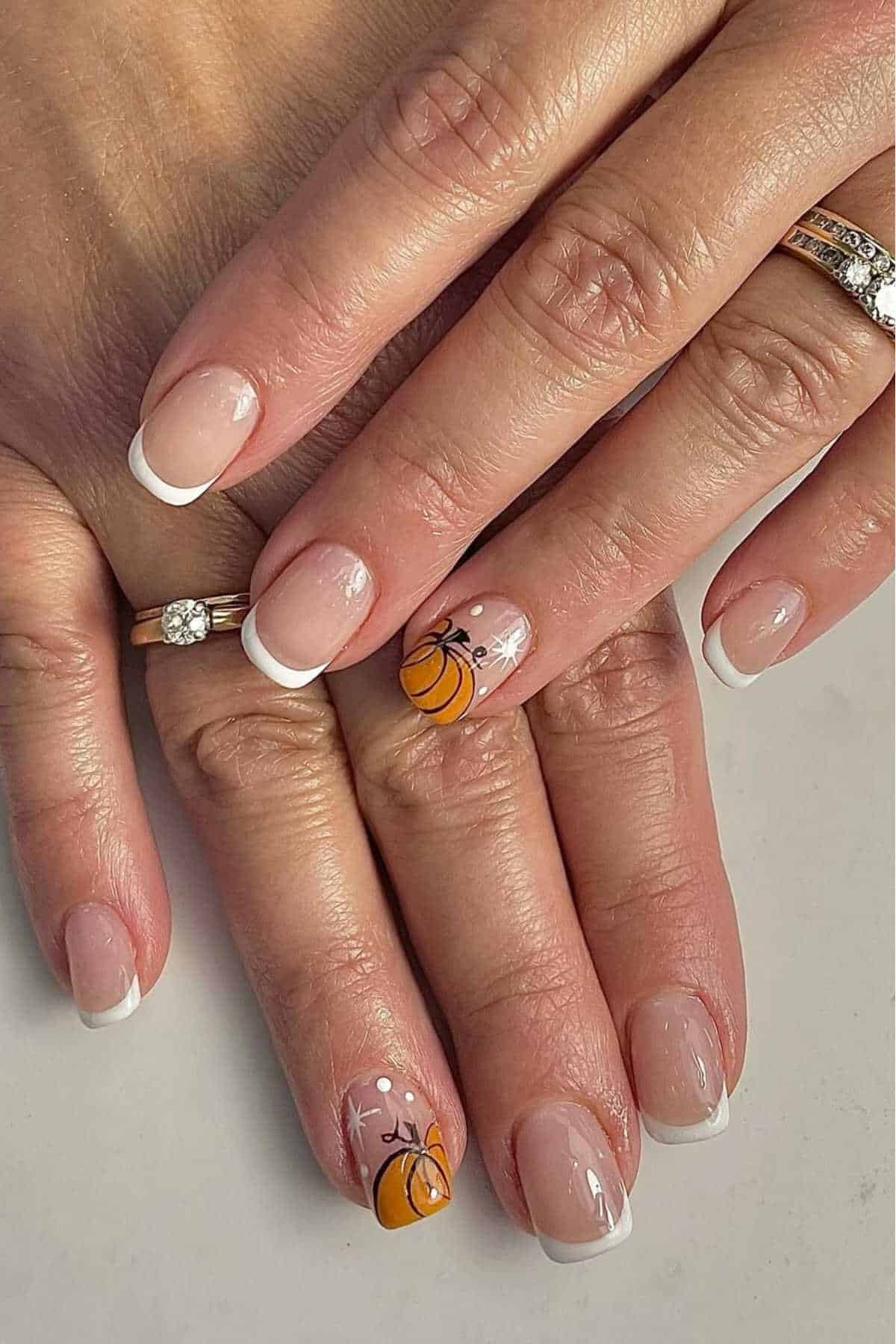 A hand with a French manicure; the ring finger nail features a small orange pumpkin and white floral design. The person wears two rings, one with a diamond.