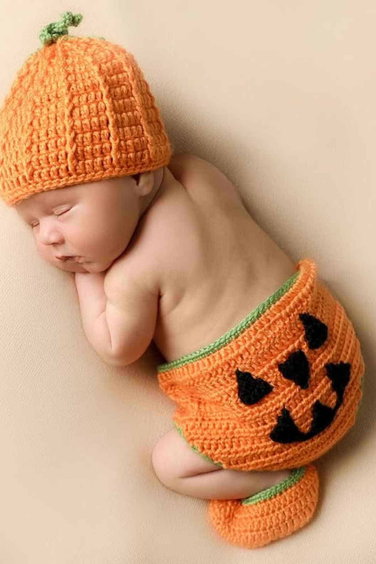 Newborn baby sleeping while wearing a crocheted orange pumpkin hat and diaper cover with black triangle patterns on a beige background.