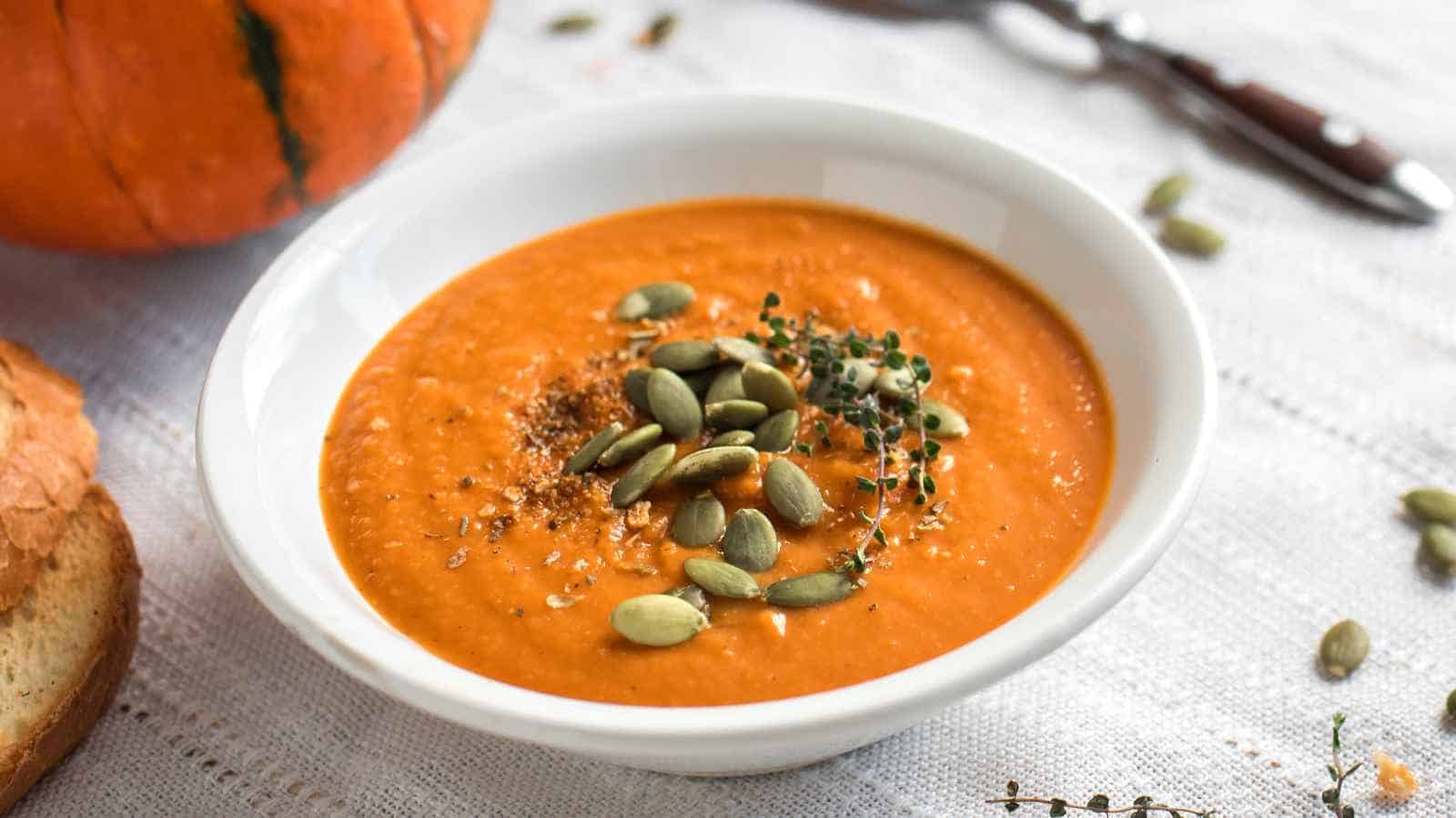 A white bowl filled with orange pumpkin soup, topped with pepitas and herbs, sits on a white tablecloth next to a slice of bread.