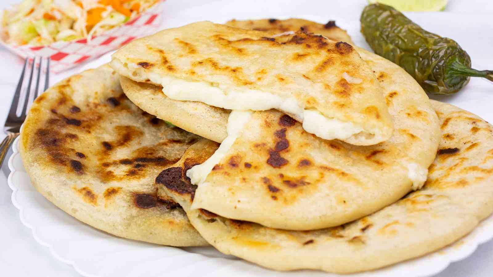 Three cheese-filled pupusas on a white plate, with one pupusa partially split open to show the melted filling. A pickled vegetable side and a green chili are in the background.