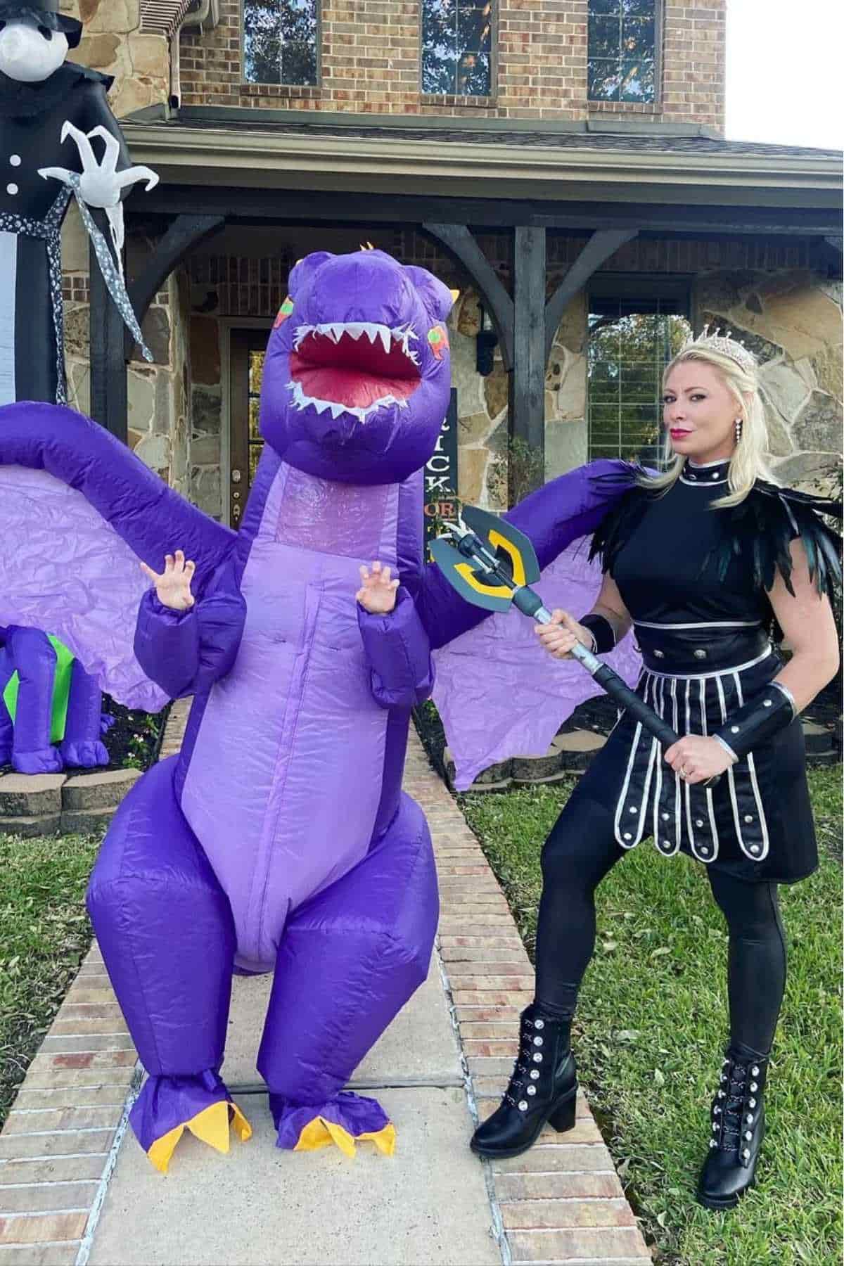 An adult in a black warrior costume and a child in an inflatable purple dragon costume stand on a walkway in front of a house decorated for Halloween.